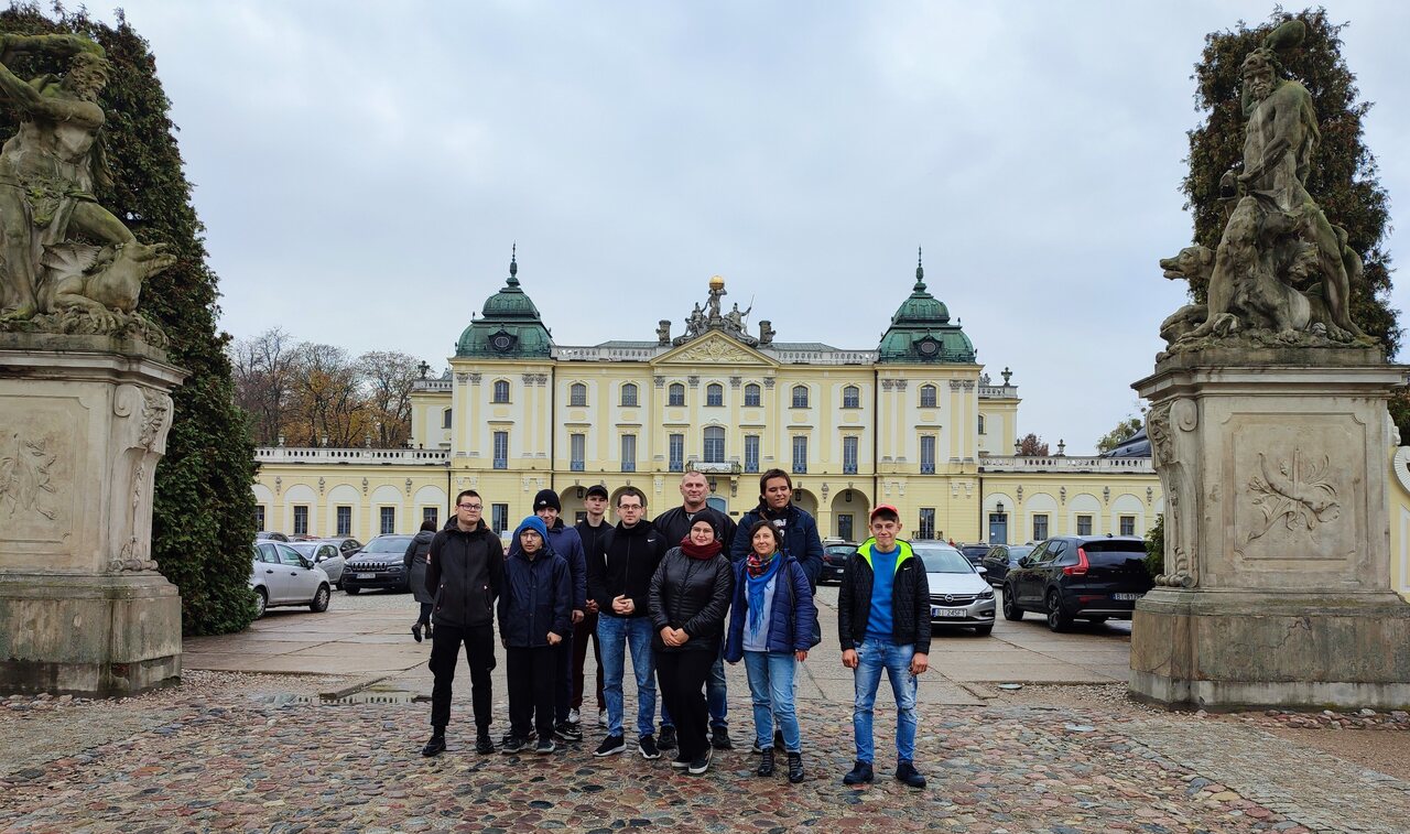 Grupa stoi na placu przed główną fasadą Pałacu Branickich, pomiędzy dwoma dużymi rzeźbami.