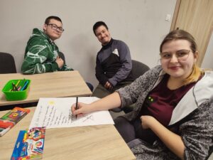 Uczestnicy siedzą przy stołach i na kanapie, wykonując prace plastyczne w asyście wychowawcy podczas warsztatów.