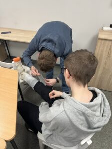 Zd.2 Młodzież uczy się, jak nieść pierwsza pomoc. Na zdjęciu kolega unieruchamia szyną i bandażem nogę drugiemu koledze