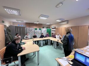 Zdj. 2 - wychowankowie siedzą przy stole, przed nimi stoi pracownica Urzędu i opowiada, o czym będzie dzisiejsze spotkanie.
