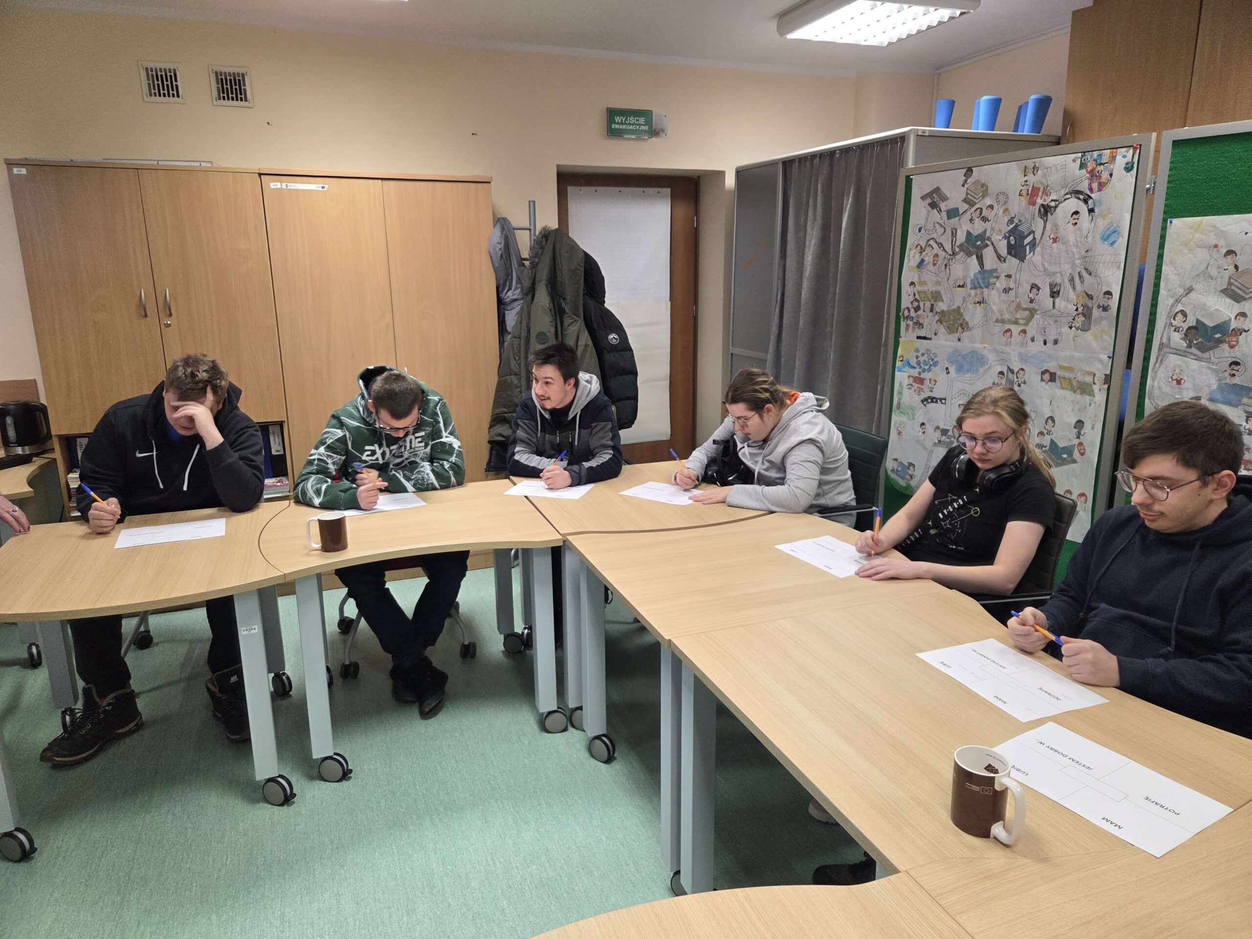 Zdj. 3 - wychowankowie siedzą przy stole, przed sobą mają kartki papieru, w rękach trzymają długopisy.
