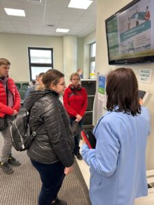 Zdj. 1
Uczniowie z Zespołu Szkół nr 16 w Białymstoku przy automacie w siedzibie ZUS. Pani w niebieskiej marynarce, prowadząca zajęcia, tłumaczy sposób pobierania biletu i obsługi systemu kolejkowego.
