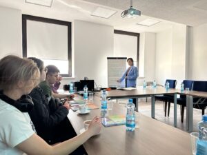 Zdj. 3
Podczas zajęć w sali konferencyjnej uczniowie z zainteresowaniem uczestniczą w spotkaniu. Prowadząca przedstawia prezentację o zadaniach ZUS i roli tej instytucji w życiu obywateli.
