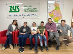 Zdj. 4
Wspólne zdjęcie pamiątkowe w siedzibie ZUS w Białymstoku. Na kanapie siedzą uczestnicy wraz z wychowawcami – blondynka po lewej stronie oraz blondyn w okularach po prawej, kucający obok grupy.
