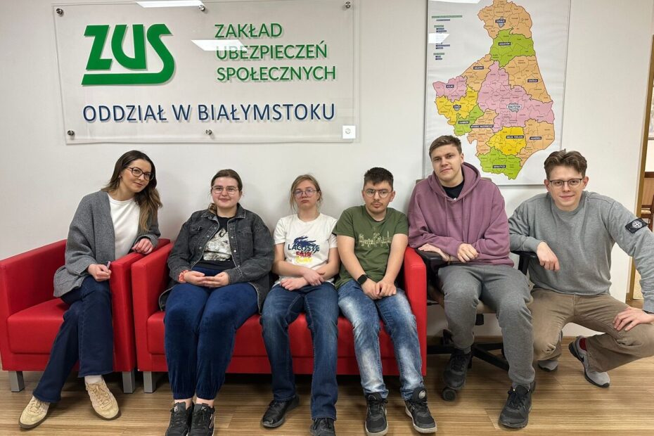 Zdj. 4 Wspólne zdjęcie pamiątkowe w siedzibie ZUS w Białymstoku. Na kanapie siedzą uczestnicy wraz z wychowawcami – blondynka po lewej stronie oraz blondyn w okularach po prawej, kucający obok grupy.