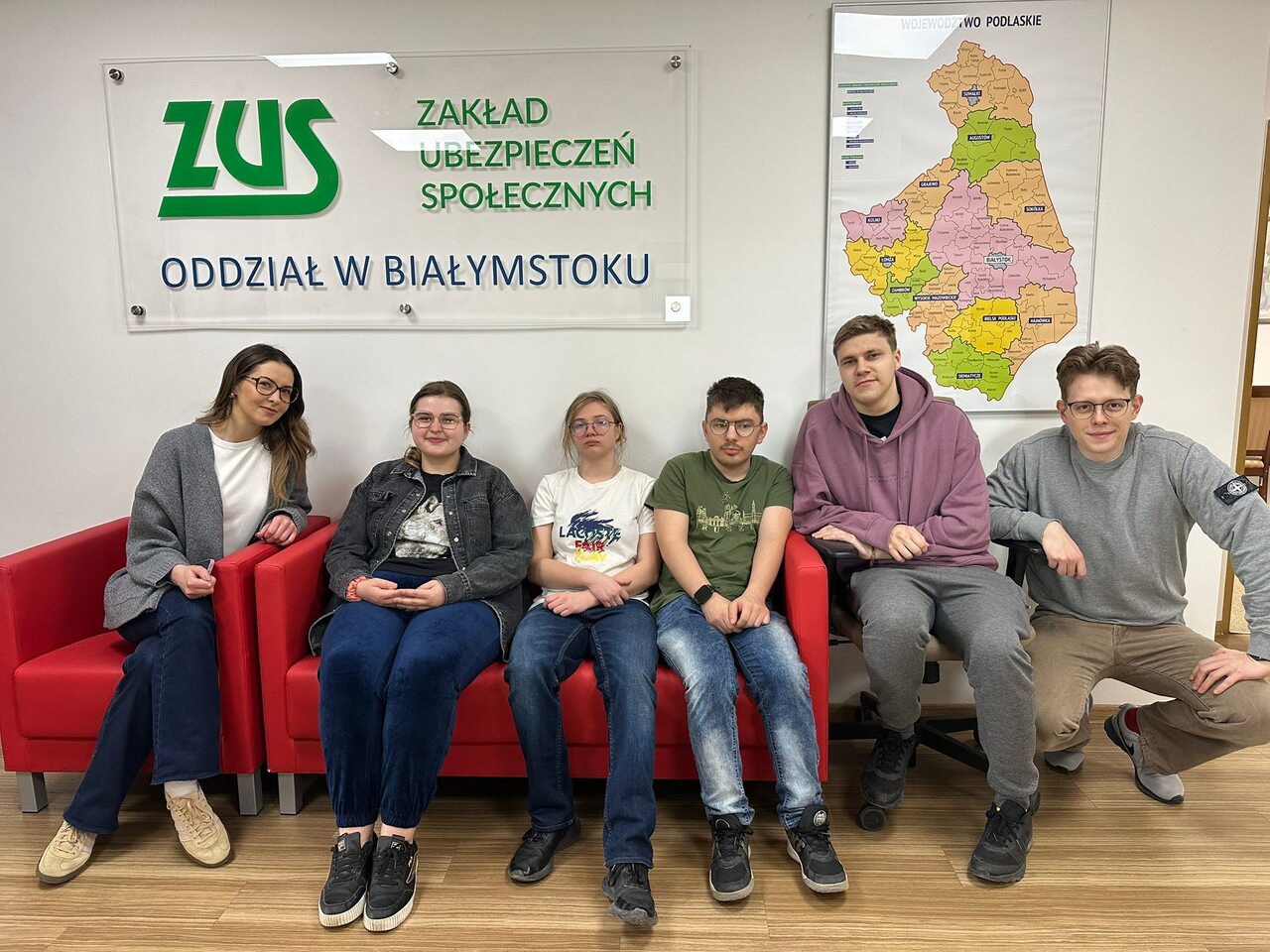 Zdj. 4
Wspólne zdjęcie pamiątkowe w siedzibie ZUS w Białymstoku. Na kanapie siedzą uczestnicy wraz z wychowawcami – blondynka po lewej stronie oraz blondyn w okularach po prawej, kucający obok grupy.
