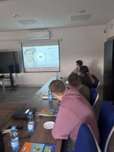 Zdj. 5
Uczniowie oglądają prezentację multimedialną o zadaniach ZUS wyświetlaną na ekranie. Osoba w okularach, siedząca na końcu stołu, to wychowawca – pozostali to uczniowie biorący udział w spotkaniu.
