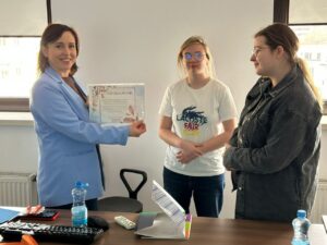 Zdj. 6
Wychowanki wręczają podziękowanie pracownicy ZUS-u.  Atmosfera spotkania jest przyjazna i pełna zaangażowania, co widać na twarzach uczestniczek.