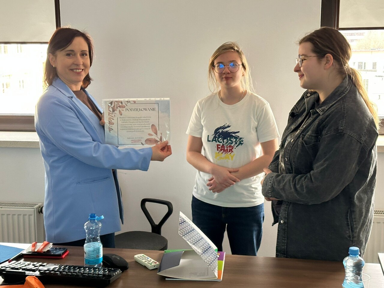 Zdj. 6
Wychowanki wręczają podziękowanie pracownicy ZUS-u.  Atmosfera spotkania jest przyjazna i pełna zaangażowania, co widać na twarzach uczestniczek.