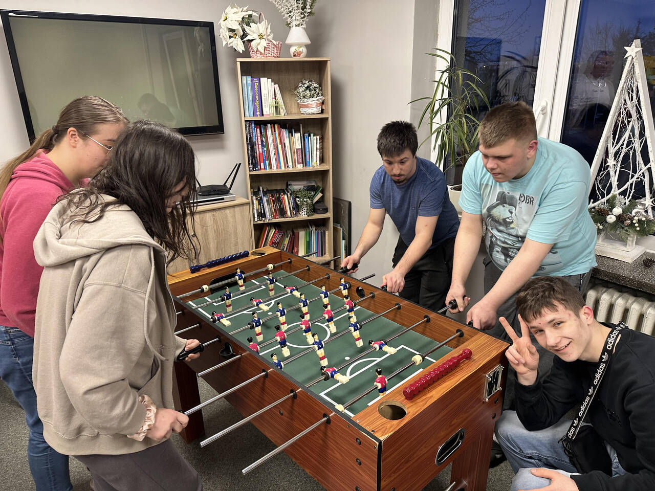 Zdjęcie 1 Pięcioro wychowanków stoi wokół drewnianego stołu do piłkarzyków w świetlicy internatu; dwie osoby po lewej, dwie po prawej, jedna kuca z przodu i pokazuje znak „V” palcami, wszyscy skupieni na grze i uśmiechnięci, w tle telewizor na ścianie, regał z książkami i okno z zimową dekoracją.