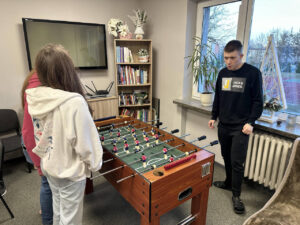 Zdjęcie 2 Na środku stoi stół do piłkarzyków, przy którego bokach grają trzy osoby, a za stołem, na tle regału z książkami i telewizora, stoi dorosły mężczyzna w jasnym T‑shircie, pełniący rolę sędziego lub opiekuna; wszyscy skoncentrowani na meczu.