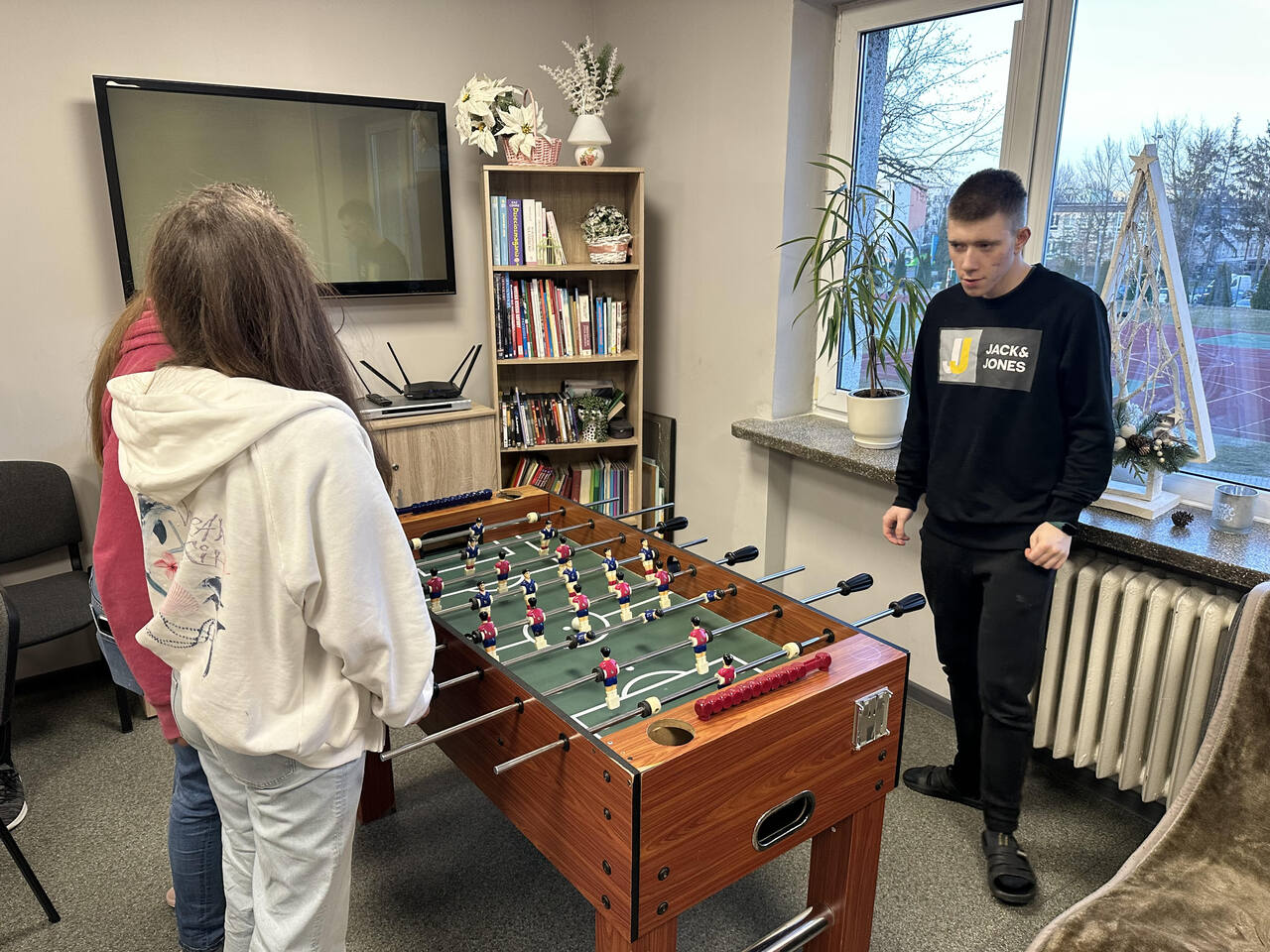 Zdjęcie 2 Na środku stoi stół do piłkarzyków, przy którego bokach grają trzy osoby, a za stołem, na tle regału z książkami i telewizora, stoi dorosły mężczyzna w jasnym T‑shircie, pełniący rolę sędziego lub opiekuna; wszyscy skoncentrowani na meczu.
