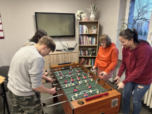 Zdjęcie 3 Przy stole do piłkarzyków stoją dwie dziewczyny po lewej stronie oraz chłopak po prawej, blisko okna z widokiem na boisko szkolne; uczestnicy czekają na rozpoczęcie gry, atmosfera jest spokojna i skupiona.