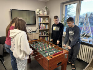 Zdjęcie 4 Cztery osoby – dwie po lewej i dwie po prawej stronie – trzymają rączki od piłkarzyków, ustawione jak do meczu; stoją w świetlicy internatu, w tle widać telewizor, regał z książkami i zimowe dekoracje w oknie.