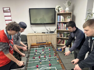 Zdjęcie 5 Cztery osoby pochylają się nad stołem do piłkarzyków w trakcie dynamicznej rozgrywki; po lewej stronie stoją dwie uczennice, po prawej dwóch uczniów, wszyscy w luźnych, kolorowych ubraniach, w tle jasna ściana, regał oraz okno z dekoracjami.