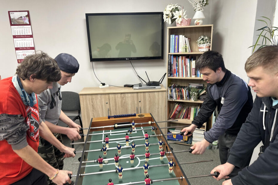 Zdjęcie 5 Cztery osoby pochylają się nad stołem do piłkarzyków w trakcie dynamicznej rozgrywki; po lewej stronie stoją dwie uczennice, po prawej dwóch uczniów, wszyscy w luźnych, kolorowych ubraniach, w tle jasna ściana, regał oraz okno z dekoracjami.