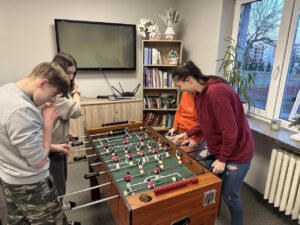 Zdjęcie 6 Czterech chłopców bierze udział w meczu piłkarzyków, stojąc po dwóch z każdej strony stołu; wszyscy mocno trzymają rączki i są skupieni na grze, za nimi widać telewizor, szafkę i regał z książkami, co podkreśla, że akcja toczy się w świetlicy internatu.