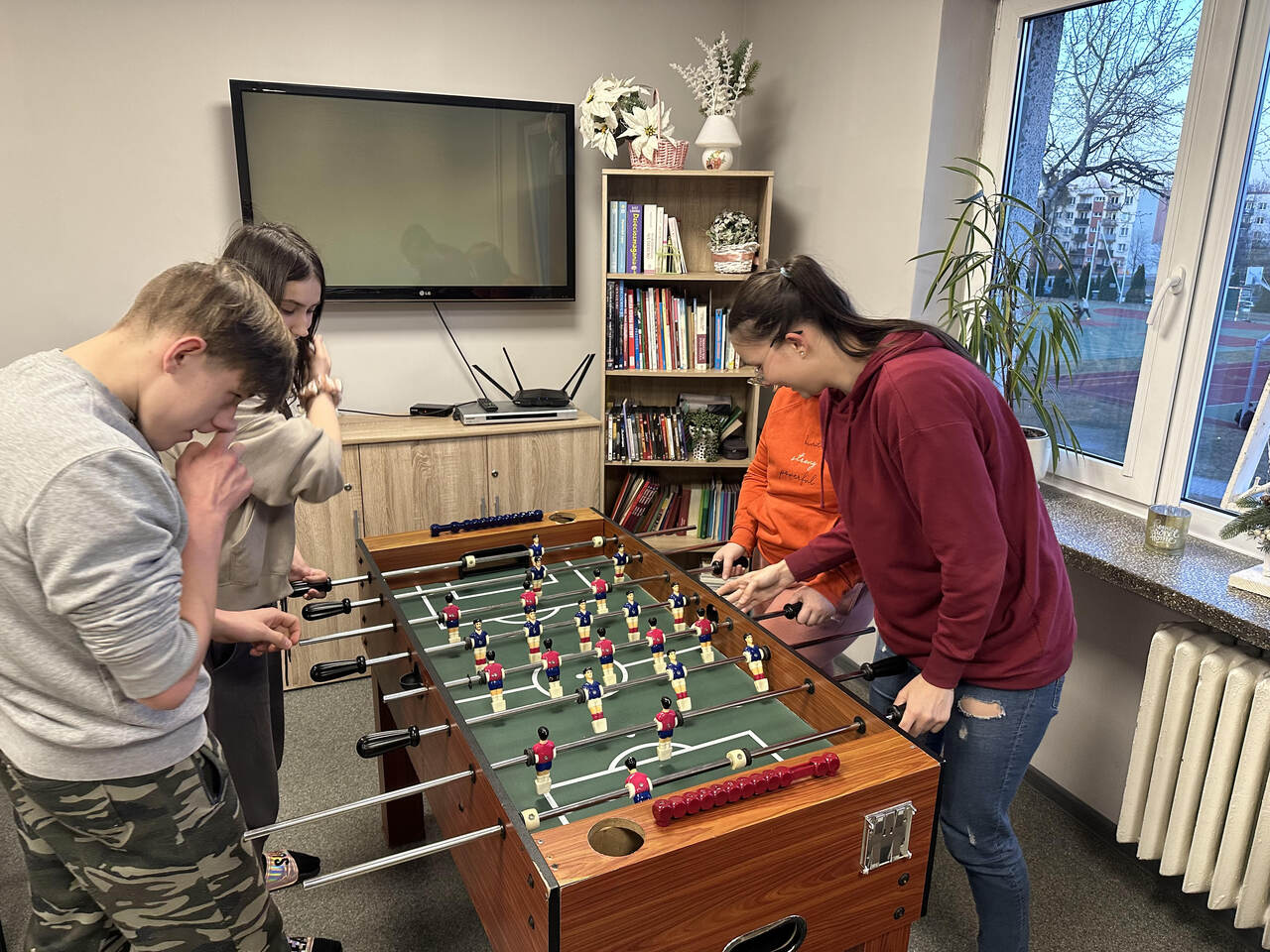 Zdjęcie 6 Czterech chłopców bierze udział w meczu piłkarzyków, stojąc po dwóch z każdej strony stołu; wszyscy mocno trzymają rączki i są skupieni na grze, za nimi widać telewizor, szafkę i regał z książkami, co podkreśla, że akcja toczy się w świetlicy internatu.