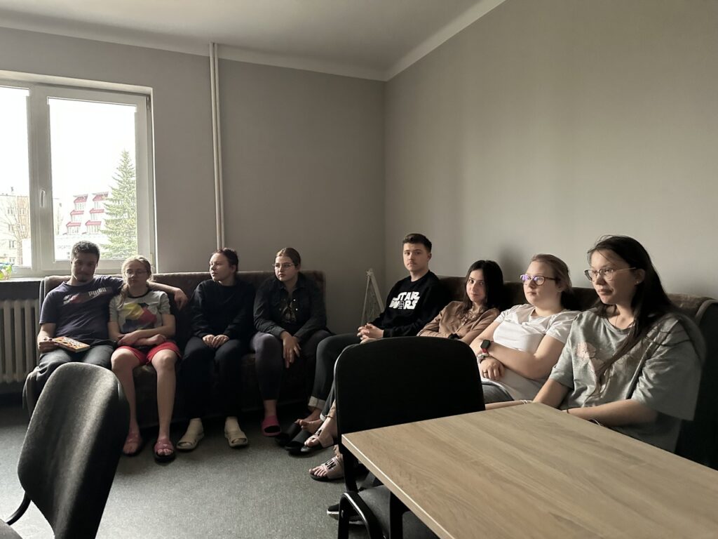 ZDJĘCIE 1
Na szarej kanapie w świetlicy internatu siedzi obok siebie ośmioosobowa grupa wychowanków; wszyscy zwróceni są w jedną stronę, jakby oglądali film, w pomieszczeniu jest półmrok, a z lewej strony widać okno z dziennym światłem.
