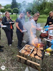 Zdjęcie 1
Na pierwszym zdjęciu widać drewniany stół ustawiony w przestrzeni przy wejściu do internatu, zastawiony miskami z truskawkami, blachami z karkówką z grilla, kawałkami kurczaka i tacą czekoladowych babeczek posypanych orzechami.
Po prawej stronie kadru stoi uczeń w czarnej koszulce z napisem „Future”, pokazujący kciuk uniesiony do góry, a w tle kilka osób nakłada sobie jedzenie na talerzyki.






