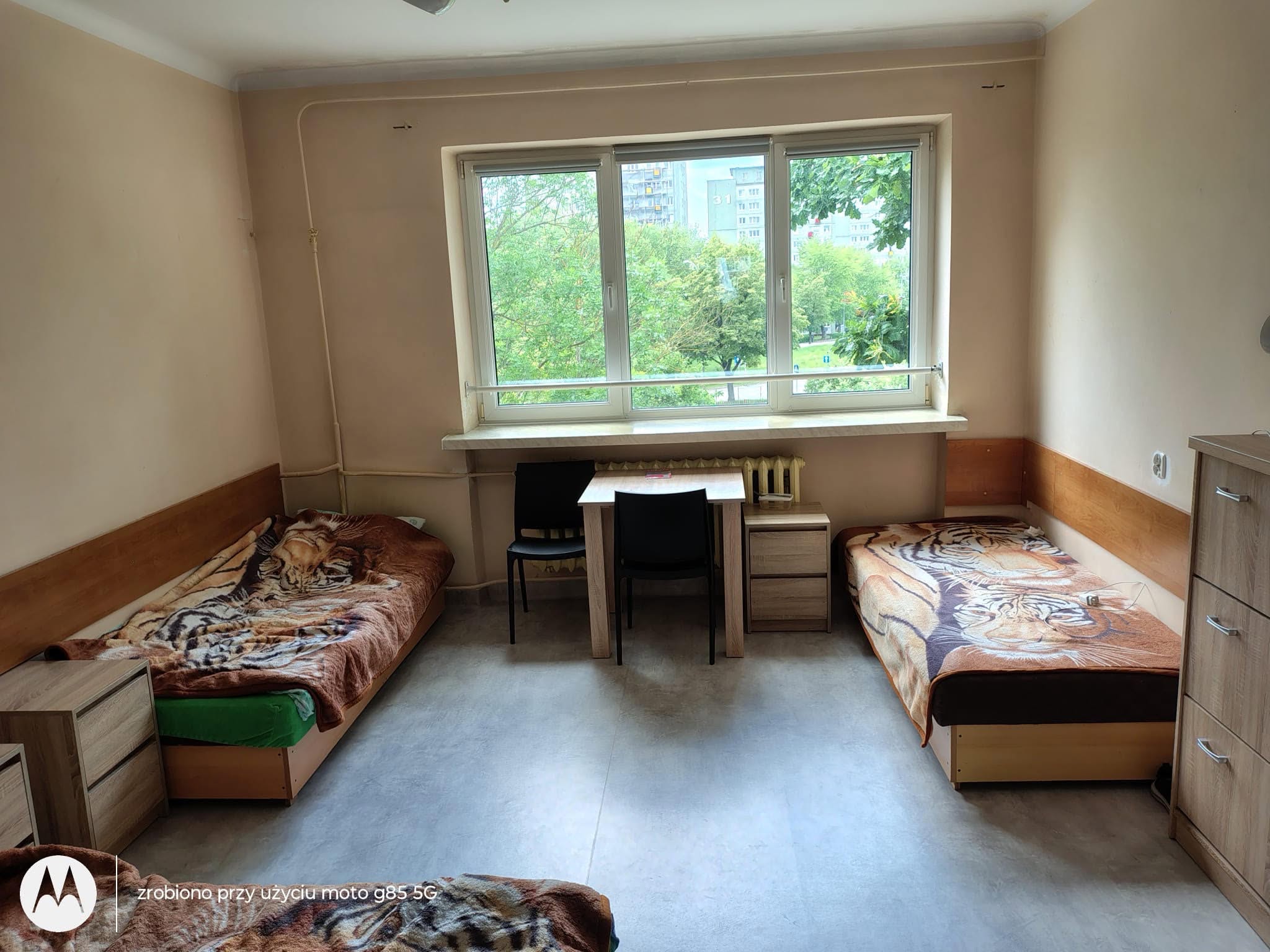 Zdjęcie 1
Uporządkowany pokój w internacie z dwoma łóżkami, stolikiem pod oknem i równo ustawionymi meblami.
Wnętrze jest czyste, schludne i przygotowane wzorowo.
