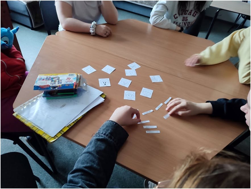 zdjęcie 10.

Grupa dzieci siedzi przy drewnianym stole i uczestniczy w grze edukacyjnej. Na blacie leżą małe karty z rysunkami twarzy przedstawiających emocje oraz paski z nazwami uczuć. Jedno z dzieci dopasowuje etykiety do odpowiednich ilustracji. Na stole znajdują się także zeszyty i przybory szkolne.
