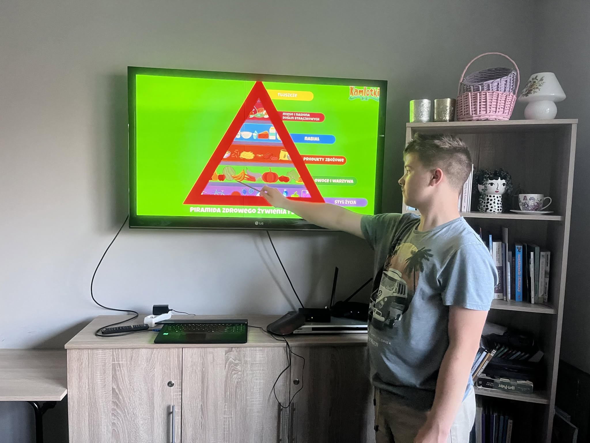 Zdj. 2 Wychowankowie stoją przy tablicy i pokazują produkty znajdujące się w piramidzie żywienia.