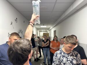 Zdjęcie 2
Grupa uczniów stoi na korytarzu internatu podczas ogłoszenia wyników konkursu.
Na pierwszym planie jedna osoba unosi nagrodę do góry, a pozostali obserwują rozstrzygnięcie.
