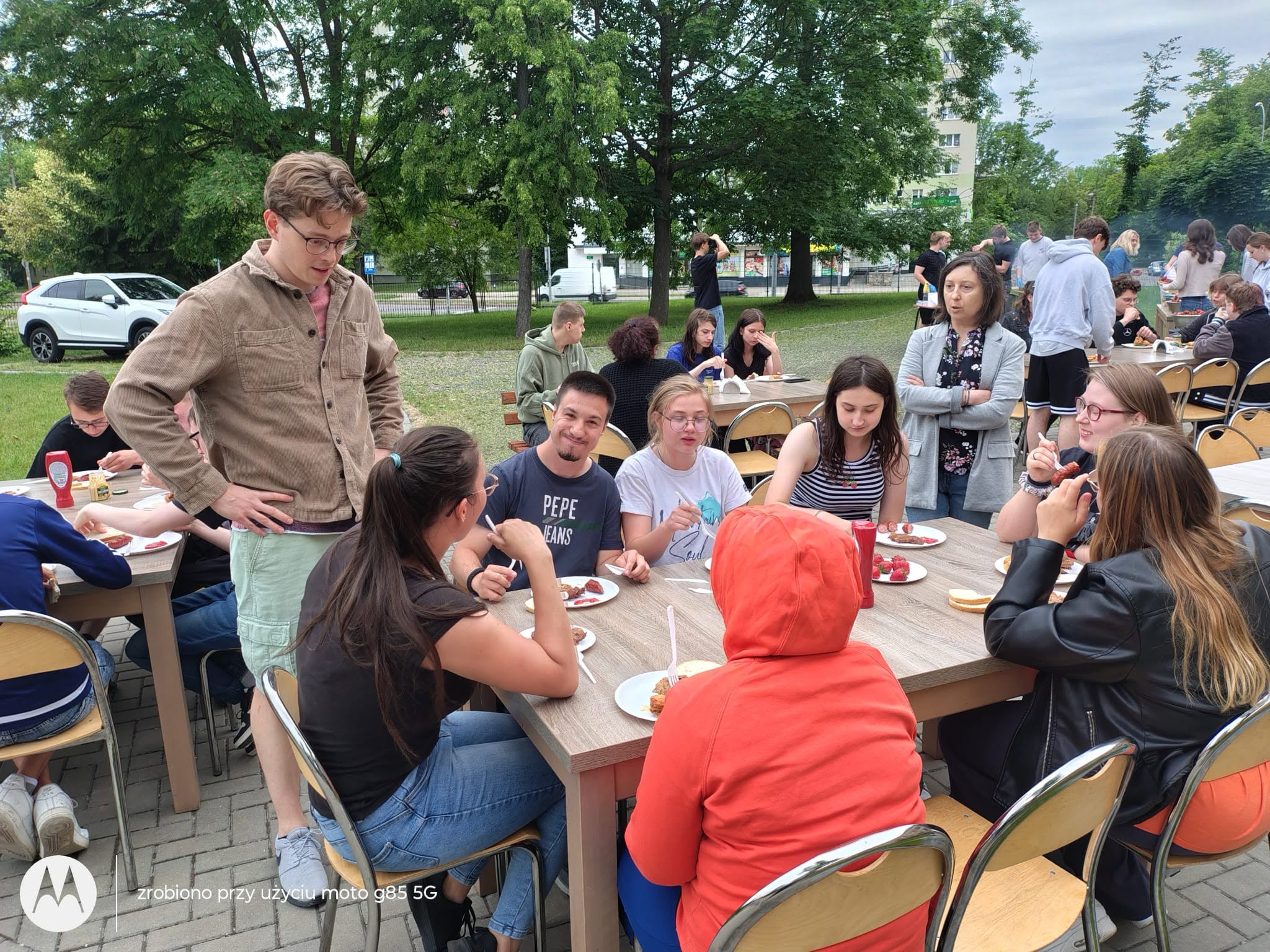 Zdjęcie 3
Na trzecim zdjęciu w centrum kadru znajduje się duży metalowy grill, na którym leżą tacki z przypieczoną karkówką oraz rzędy podsmażonych kiełbasek, nad którymi unosi się płomień i gęsty dym.
Przed grillem stoi kilka osób z plastikowymi talerzykami w dłoniach, ustawionych w luźnej kolejce, a po prawej stronie widać osobę w niebieskich rękawiczkach, która szczypcami nakłada kiełbasę na talerze.
