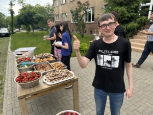 Zdjęcie 4
Czwarte zdjęcie pokazuje stół ustawiony w przestrzeni przy wejściu do internatu, przy którym siedzi grupa uczniów i osób dorosłych, jedzących potrawy z grilla i truskawki z jednorazowych talerzyków.
Przy stole widać kilka osób wykonujących gest „pokój” dwoma palcami w stronę aparatu, a w tle rozciąga się zielona okolica z drzewami i zabudowaniami.