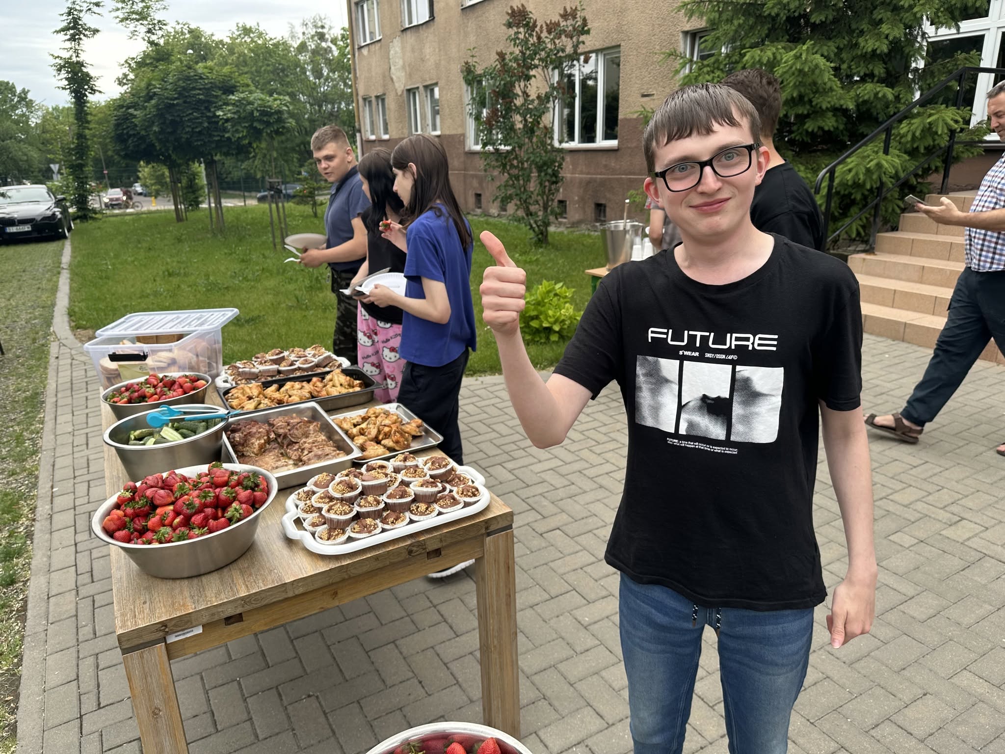 Zdjęcie 4
Czwarte zdjęcie pokazuje stół ustawiony w przestrzeni przy wejściu do internatu, przy którym siedzi grupa uczniów i osób dorosłych, jedzących potrawy z grilla i truskawki z jednorazowych talerzyków.
Przy stole widać kilka osób wykonujących gest „pokój” dwoma palcami w stronę aparatu, a w tle rozciąga się zielona okolica z drzewami i zabudowaniami.